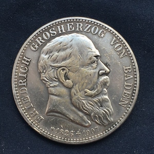 May include: Silver coin with a profile portrait of a man with a beard. The coin reads "GROSSHERZOG VON BADEN" and "FRIEDRICH *1826 +1907".