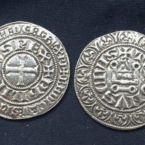 Può includere: Due monete rotonde color argento con intricati disegni in rilievo. Una moneta presenta una croce al centro, mentre l'altra ha un motivo complesso. Le monete sono poste su uno sfondo blu scuro.