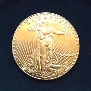 May include: A gold-colored coin with a depiction of Liberty, a woman holding a torch and an olive branch, standing on a rock with a sunburst design behind her. The inscription "LIBERTY" is visible at the top of the coin.