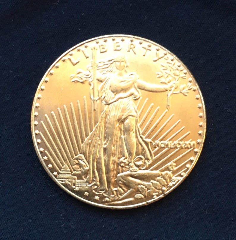 May include: A gold-colored coin with a depiction of Liberty, a woman holding a torch and an olive branch, standing on a rock with a sunburst design behind her. The inscription "LIBERTY" is visible at the top of the coin.