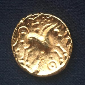 May include: A gold coin with an embossed design featuring a horse-like creature. The coin has a worn appearance, with visible imperfections and a slightly irregular shape. The background is a dark blue.