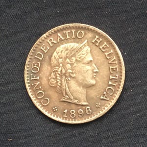 Op de afbeelding: Een bronzen munt uit 1896 met de inscriptie "CONFOEDERATIO HELVETICA". De munt toont een profiel van een vrouw met een lauwerkrans. De munt heeft een getextureerde rand. Donkere achtergrond.