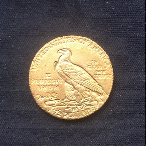 May include: A gold-coloured coin with the inscription "United States of America" and "In God We Trust" around the edge. The centre of the coin features an eagle with outstretched wings and the inscription "E Pluribus Unum" and "2 1/2 Dollars".