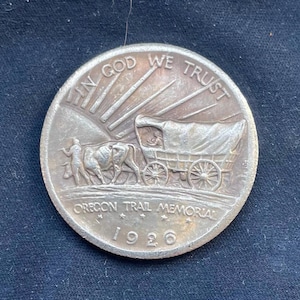 Può includere: Una moneta d'argento del Memoriale dell'Oregon Trail del 1926. La moneta presenta un carro coperto trainato da buoi, una figura e l'iscrizione "IN GOD WE TRUST". Sono visibili anche le parole "OREGON TRAIL MEMORIAL" e l'anno 1926.