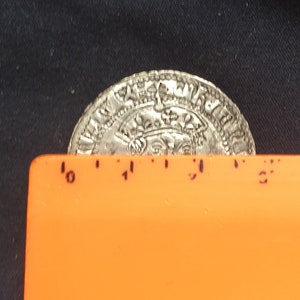 May include: A silver coin with intricate designs and lettering, measuring approximately 2.5 inches in diameter. The coin is resting on a bright orange ruler.