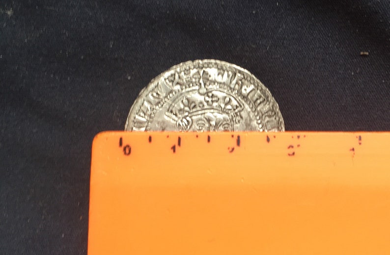 May include: A silver coin with intricate designs and lettering, measuring approximately 2.5 inches in diameter. The coin is resting on a bright orange ruler.