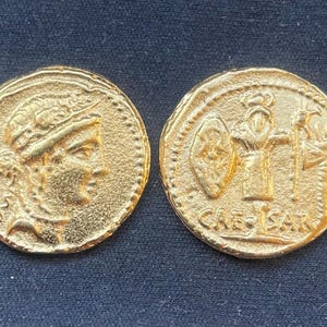 May include: Two gold-colored coins with detailed relief carvings. One coin features a profile of a person wearing a laurel wreath. The other coin has a shield, a figure, and the inscription "CAE-SAR".