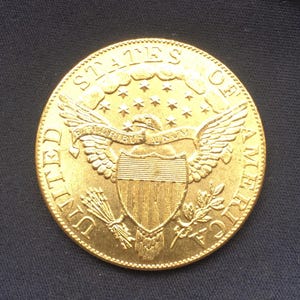 May include: A gold coin with the words "UNITED STATES OF AMERICA" around the edge. The coin features an eagle with outstretched wings, a shield, and the inscription "E PLURIBUS UNUM". The coin has a detailed design and a shiny finish.
