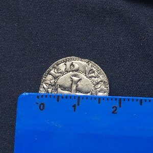 May include: A silver coin with a design of a crown and the letters "I D E" around the edge. The coin is approximately 2.5 cm in diameter.