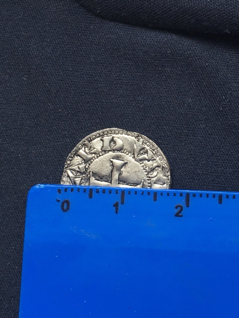 May include: A silver coin with a design of a crown and the letters "I D E" around the edge. The coin is approximately 2.5 cm in diameter.