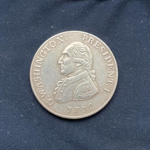 Może przedstawiać: Srebrna moneta z profilem George'a Washingtona, z napisem "G. WASHINGTON. PRESIDENT. I." i datą "1792". Moneta jest okrągła i ma szczegółowy relief.