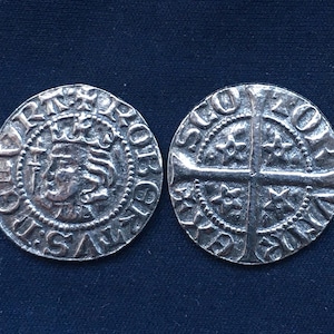 May include: Two antique silver coins with detailed engravings. One coin features a crowned profile and text around the edge. The other coin has a cross design with floral elements and text. Both coins are on a dark blue background.