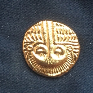 Puede incluir: Una moneda de oro con un diseño detallado de una cara con una corona y una letra 'R' en la parte inferior.