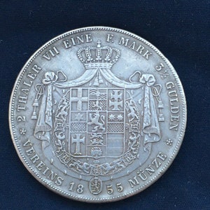 May include: A silver coin with the text "VII EINE F. MARK 5 1/2 GULDEN" and "VEREINS 18 55 MÜNZE" around the edge. The coin features a coat of arms with a crown above it.