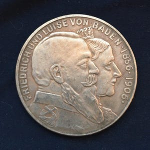May include: A silver-colored commemorative coin featuring profiles of two individuals. The coin's edge includes the text "FRIEDRICH UND LUISE VON BADEN 1856-1906". The coin has a dark background.