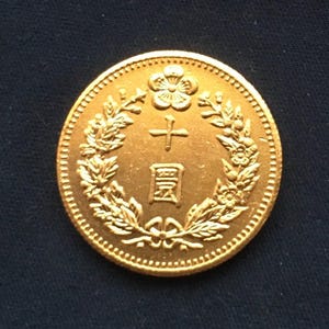 Op de afbeelding: Een gouden munt met ingewikkelde details. De munt heeft een bloemmotief aan de bovenkant, Chinese karakters in het midden en een laurierkrans rond de tekst. De rand van de munt heeft een gestructureerd patroon.