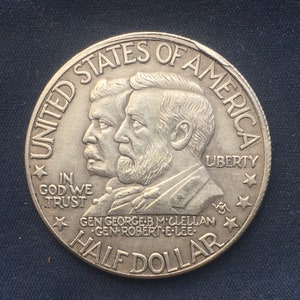 May include: A silver half dollar coin featuring the profiles of General George B. McClellan and General Robert E. Lee. The coin has the inscription "United States of America" around the rim, "In God We Trust" and "Liberty" on the obverse, and "Half Dollar" on the reverse.