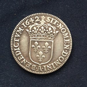 May include: A silver coin from 1642, featuring a crown and a shield with three fleur-de-lis. The coin has a textured edge and inscriptions around the border. The design is centered on a dark background.