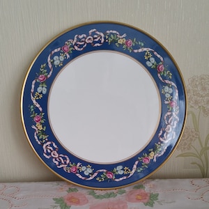 May include: A blue and white ceramic plate with a gold rim and a floral design. The plate features pink and yellow flowers with green leaves and pink ribbons.