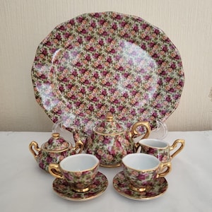 Könnte beinhalten: Ein florales Teeservice mit Teekanne, Milchkännchen, Zuckerdose, zwei Tassen und Untertassen sowie einem großen Servierteller. Das Set ist mit einem rosa-grünen Blumenmuster und goldenen Verzierungen verziert.