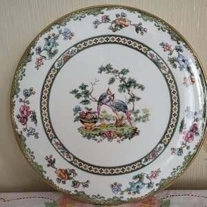 May include: A white ceramic plate with a gold rim and a floral design featuring two birds and a basket of fruit. The plate has a green and gold border with a repeating pattern.