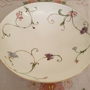 May include: A white ceramic bowl with a gold rim and a floral design. The design features pink, blue, and purple flowers with green vines.