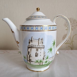 Op de afbeelding: Witte porseleinen theepot met een gouden rand en een geschilderd tafereel van een kasteel en palmbomen. De theepot heeft een deksel met een gouden knop.