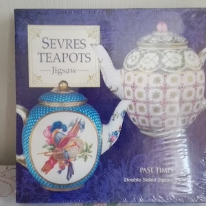 Könnte beinhalten: Eine Schachtel für ein doppelseitiges Puzzle mit zwei Sèvres-Teekannen. Die Teekannen sind beide weiß mit aufwendigen Blumendesigns. Der Text auf der Schachtel lautet "Sevres Teapots - Jigsaw - Past Times Double Sided Jigsaw Puzzle."