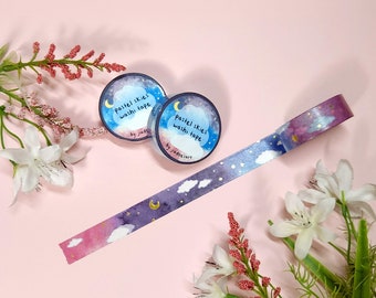 Può includere: Due rotoli di washi tape color pastello blu e viola con dettagli dorati e un motivo di nuvole bianche e stelle. Il nastro è etichettato "Pastel Skies Washi Tape" da "jua does art".