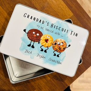 May include: A white metal tin with a blue watercolour background and the text "Grandad's Biscuit Tin To be shared with..."  Three cartoon biscuits with faces are lined up with the names Jack, Dylan, and Sebastian written below them.