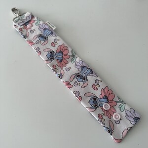 May include: A white lanyard featuring a repeating pattern of Stitch, flowers, and mushrooms in pink, blue, and green. It has a silver clasp and two pink snap fasteners.