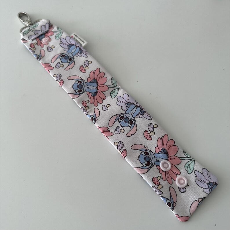 May include: A white lanyard featuring a repeating pattern of Stitch, flowers, and mushrooms in pink, blue, and green. It has a silver clasp and two pink snap fasteners.
