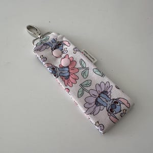 May include: A fabric keychain case with a cartoon character and floral design in pink, purple, and blue. It has a silver clasp and two pink buttons. The text "Handmade by SugarGum Designs" is visible on the label.