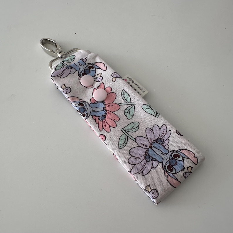May include: A fabric keychain case with a cartoon character and floral design in pink, purple, and blue. It has a silver clasp and two pink buttons. The text "Handmade by SugarGum Designs" is visible on the label.