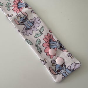May include: A light-coloured fabric headband with a repeating pattern of a blue cartoon character, pink and purple flowers, and mushrooms. Two small, light pink buttons are visible on the headband.