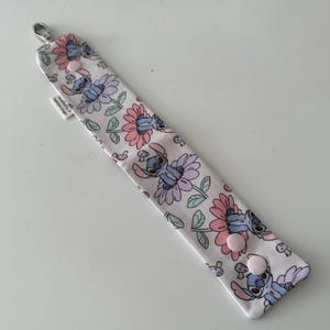 May include: A white fabric wristlet featuring a repeating pattern of Stitch from Lilo & Stitch, flowers, and mushrooms. The wristlet has a silver clasp and two pink snap closures. A small white tag is attached.