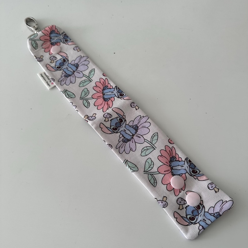 May include: A white fabric wristlet featuring a repeating pattern of Stitch from Lilo & Stitch, flowers, and mushrooms. The wristlet has a silver clasp and two pink snap closures. A small white tag is attached.