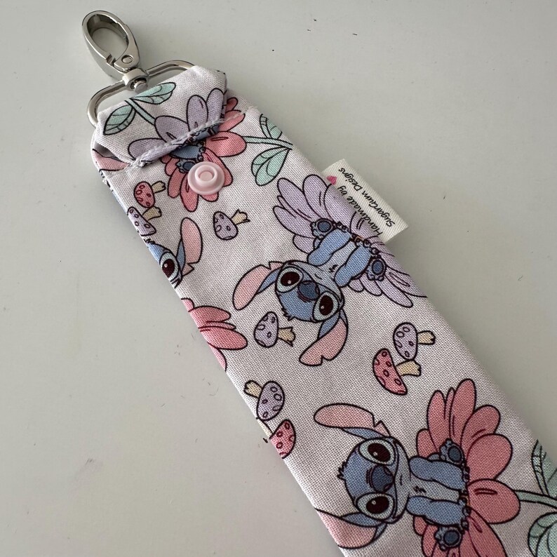May include: A light pink fabric keychain holder with a cartoon character and floral pattern. The design features a blue alien character, pink flowers, and mushrooms. It has a silver clasp and a pink button closure.