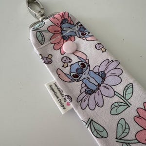 May include: A fabric case with a cartoon character design. The case features a floral pattern with a blue cartoon character, pink flowers, and green leaves. It has a pink button closure and a silver clasp. The tag reads "Handmade by SugarGum Designs."