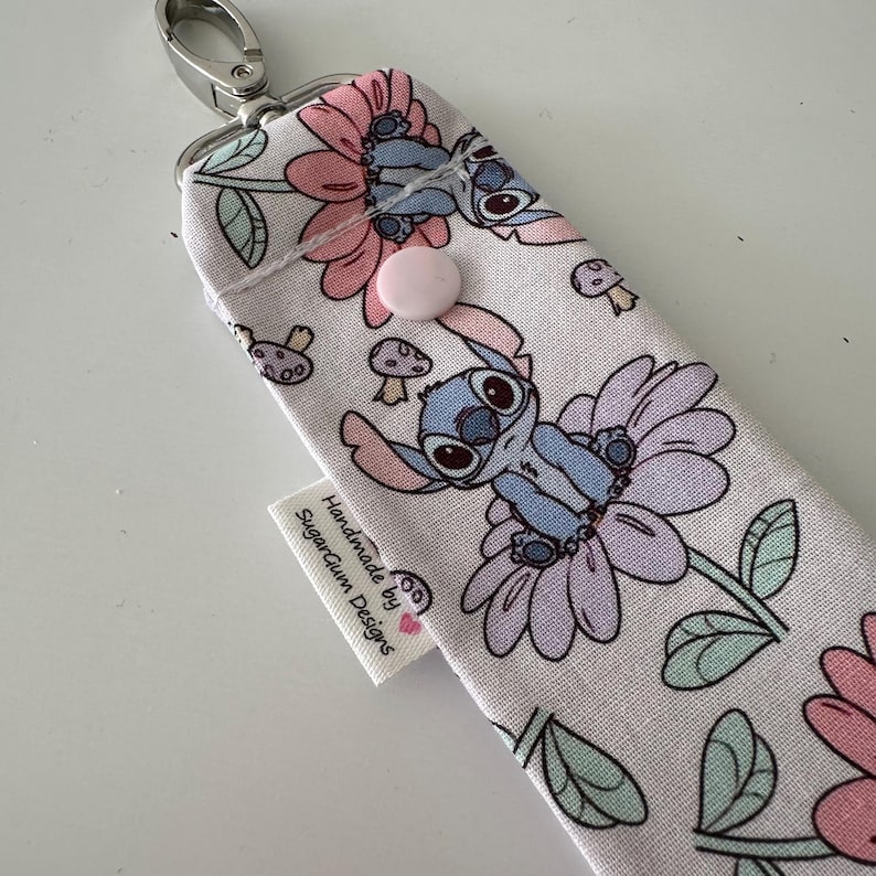 May include: A fabric case with a cartoon character design. The case features a floral pattern with a blue cartoon character, pink flowers, and green leaves. It has a pink button closure and a silver clasp. The tag reads "Handmade by SugarGum Designs."