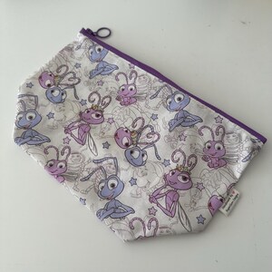 Può includere: Un astuccio bianco con cerniera con un motivo ripetuto di insetti dei cartoni animati in viola e blu. L'astuccio ha una cerniera e un bordo viola. Il design include stelle e contorni di altri personaggi. L'astuccio è etichettato "Handmade by SuperGum Designs."