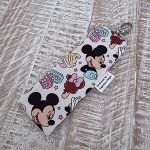 May include: A rectangular fabric pouch featuring a repeating pattern of Mickey Mouse, ice cream treats, and the word magic in blue text. The pouch has a black snap button closure, a silver metal swivel clasp, and a white tag reading Handmade by SugarGum Designs.