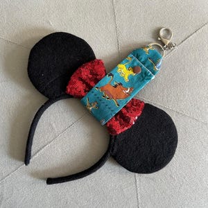 May include: Black mouse ear headband with red sequin bow details. A teal fabric keychain holder featuring characters from The Lion King is attached. The keychain holder has a silver clasp.