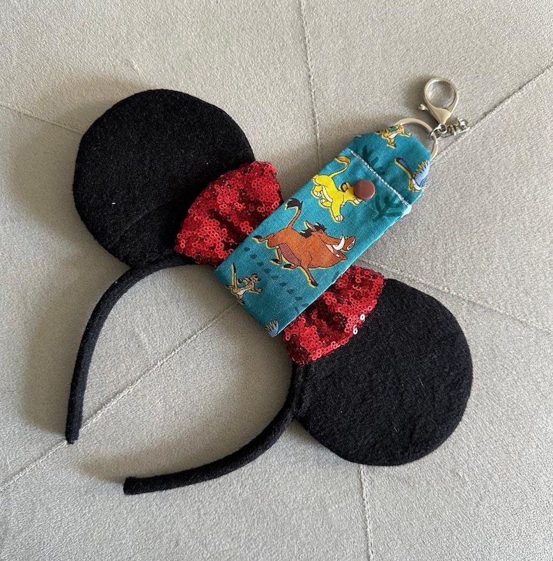 May include: Black mouse ear headband with red sequin bow details. A teal fabric keychain holder featuring characters from The Lion King is attached. The keychain holder has a silver clasp.