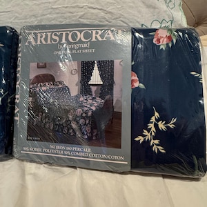 May include: A navy blue flat sheet in plastic packaging featuring a floral pattern with pink roses and green leaves. The label reads Aristocrat by Springmaid, One Full Flat Sheet, No Iron 180 Percale, 50% Kodel Polyester 50% Combed Cotton.