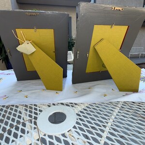 Puede incluir: Dos marcos de fotos rectangulares vistos desde atr&aacute;s, con bordes de madera pintados de gris y paneles traseros amarillos. Cada marco incluye un soporte de caballete abatible, herrajes met&aacute;licos para colgar y pesta&ntilde;as de metal.