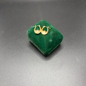 May include: A pair of small, gold-colored hoop earrings with a textured design. The earrings are displayed on a small, green velvet jewelry box against a dark background. The earrings have a classic, elegant style.