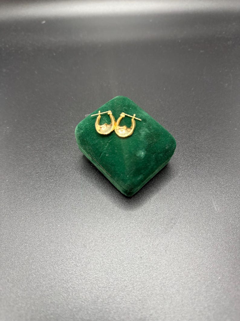 May include: A pair of small, gold-colored hoop earrings with a textured design. The earrings are displayed on a small, green velvet jewelry box against a dark background. The earrings have a classic, elegant style.