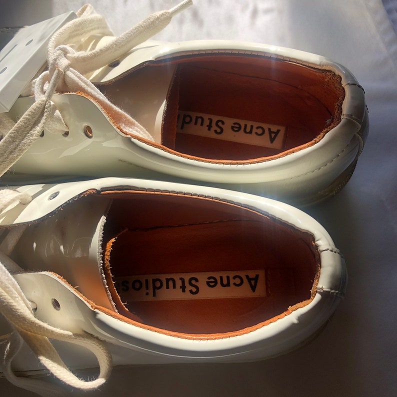 May include: A pair of white leather sneakers with white laces. The inside of the shoes are lined with brown leather and have the text "Acne Studios" printed on the insole.