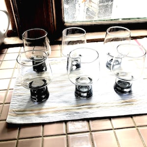Può includere: Sei bicchieri da bere in vetro trasparente con base nera sono disposti su un asciugamano da cucina a strisce bianche e marroni. I bicchieri sono su un bancone piastrellato.